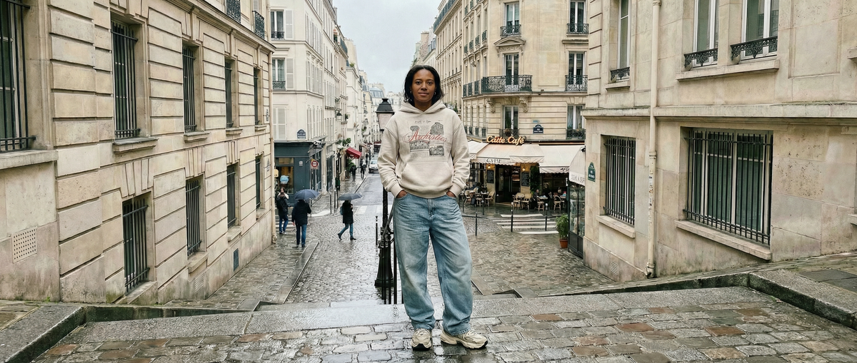 A person wearing a light-colored hoodie and loose jeans stands confidently on a cobblestone street flanked by classic European-style buildings, with a few pedestrians in the background.