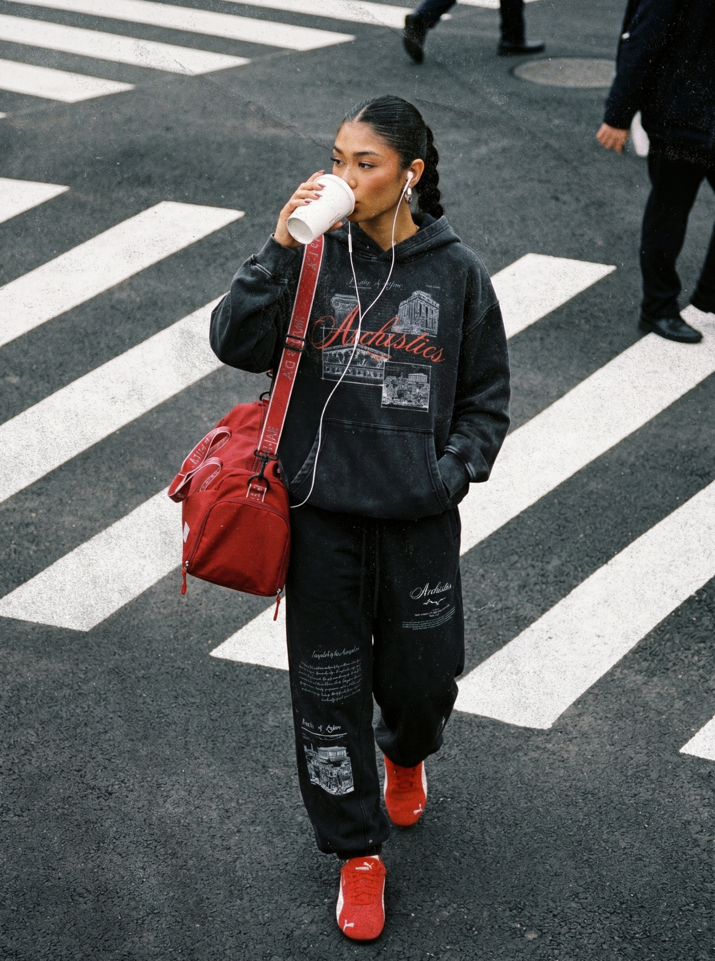 Person crossing a zebra crossing, wearing a black hoodie and pants with red shoes and bag.