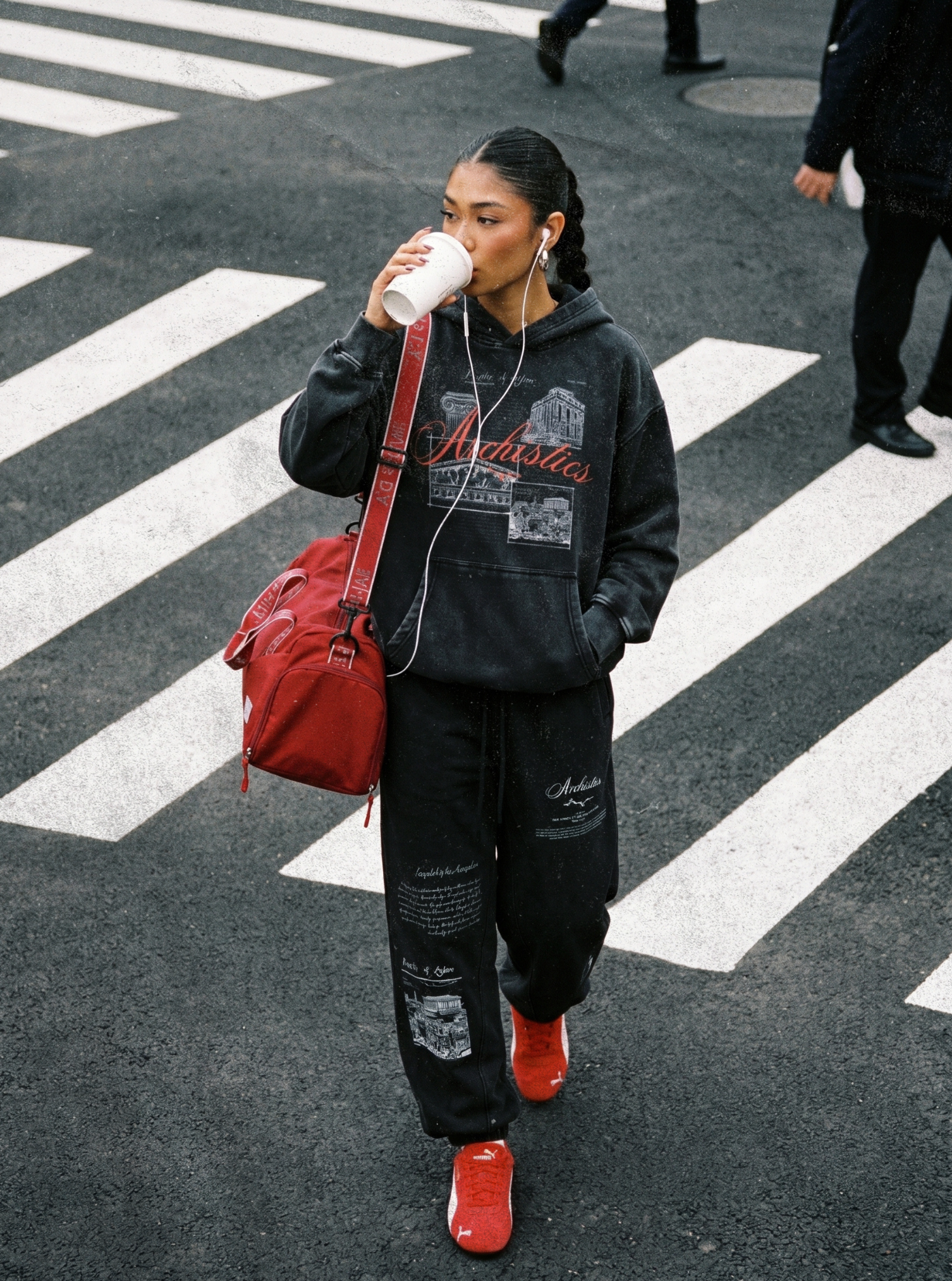 Person crossing a zebra crossing, wearing a black hoodie and pants with red shoes and bag.