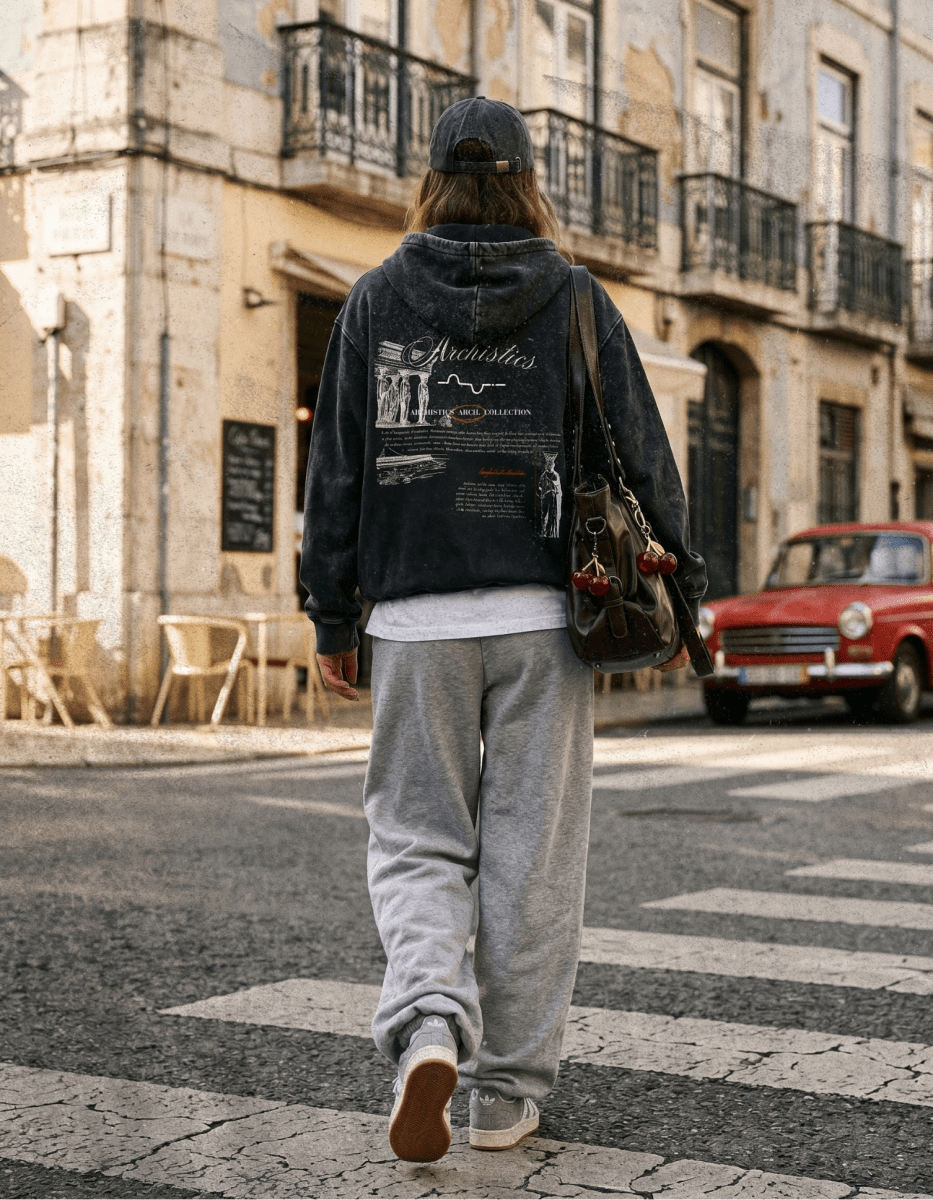 Person walking on a street wearing a black hoodie with text and graphics, gray sweatpants, and a cap.