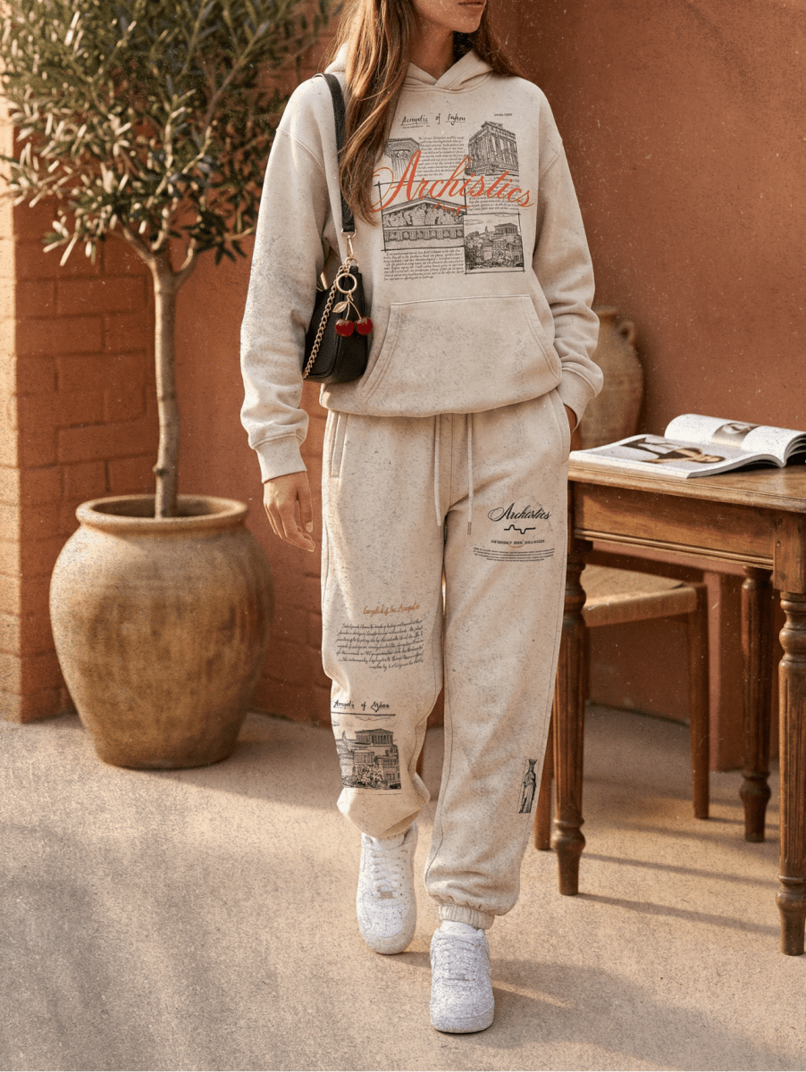Person wearing a beige hoodie and jogger set with 'Antarctica' text, standing in front of a rustic wall.