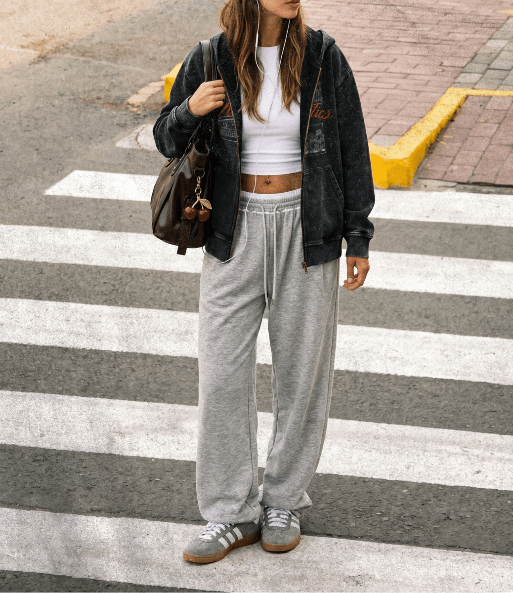 Person wearing a black jacket, white crop top, gray sweatpants, and Adidas shoes on a crosswalk.