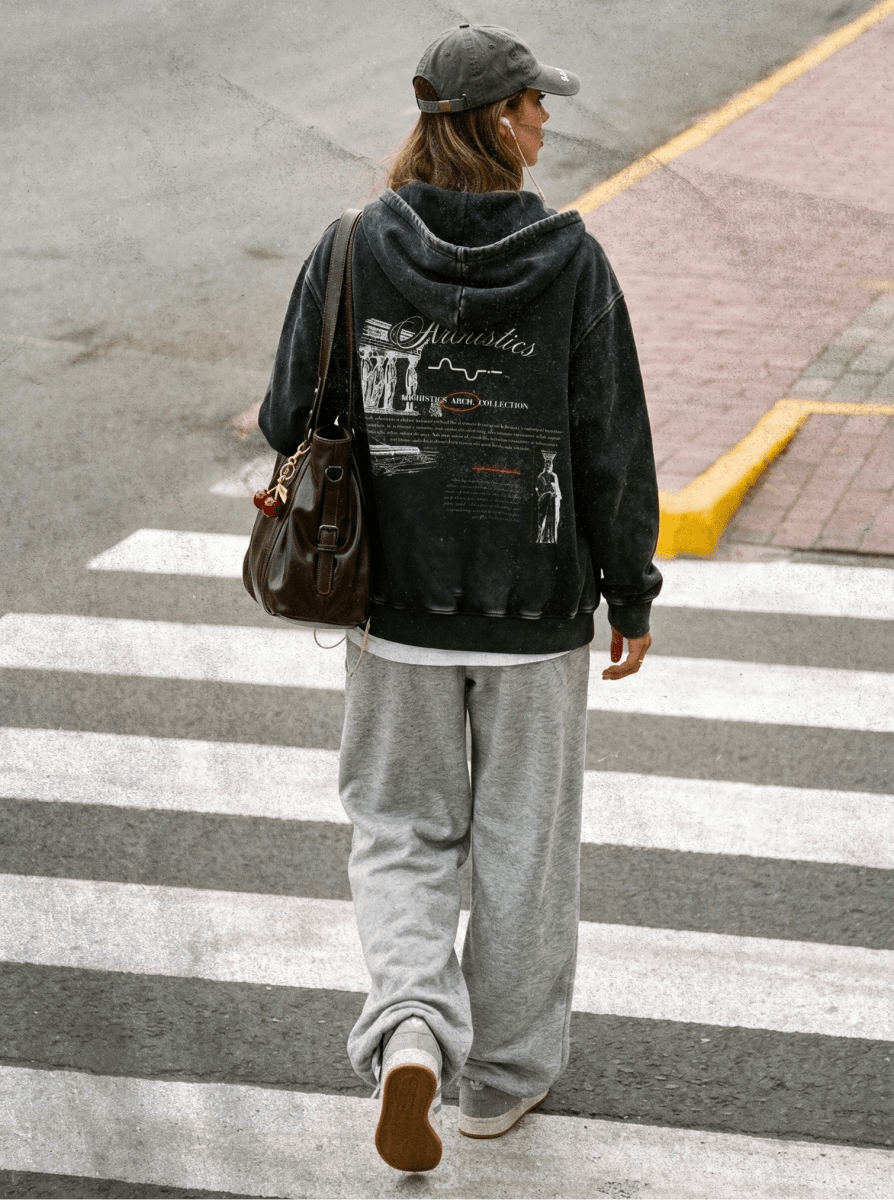 Person wearing a black hoodie with text and graphics, gray pants, and a cap, crossing a street.