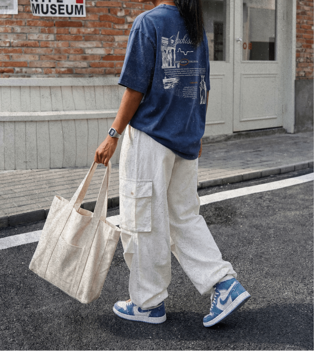 Person wearing a blue t-shirt and white pants, holding a beige tote bag, walking on a street.