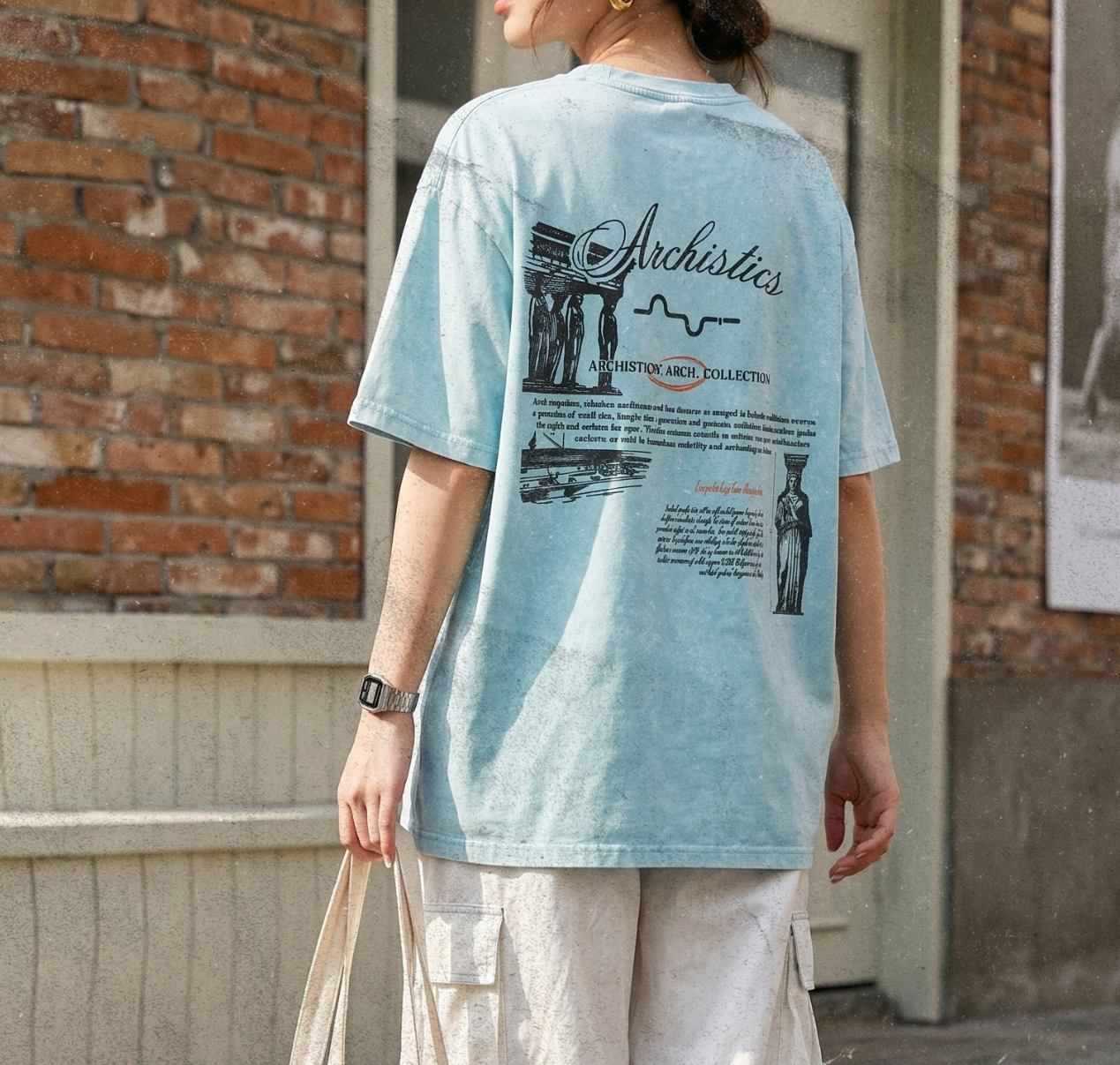 Person wearing a light blue t-shirt with text and graphics, holding a beige tote bag, standing on a street.