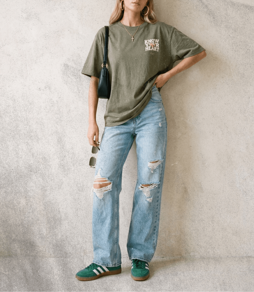 Person wearing an olive green t-shirt and ripped jeans against a textured wall.