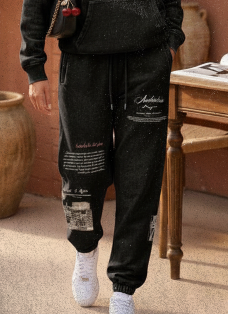 Woman in black hoodie and pants standing in a warm-toned outdoor setting with a plant and table in the background.